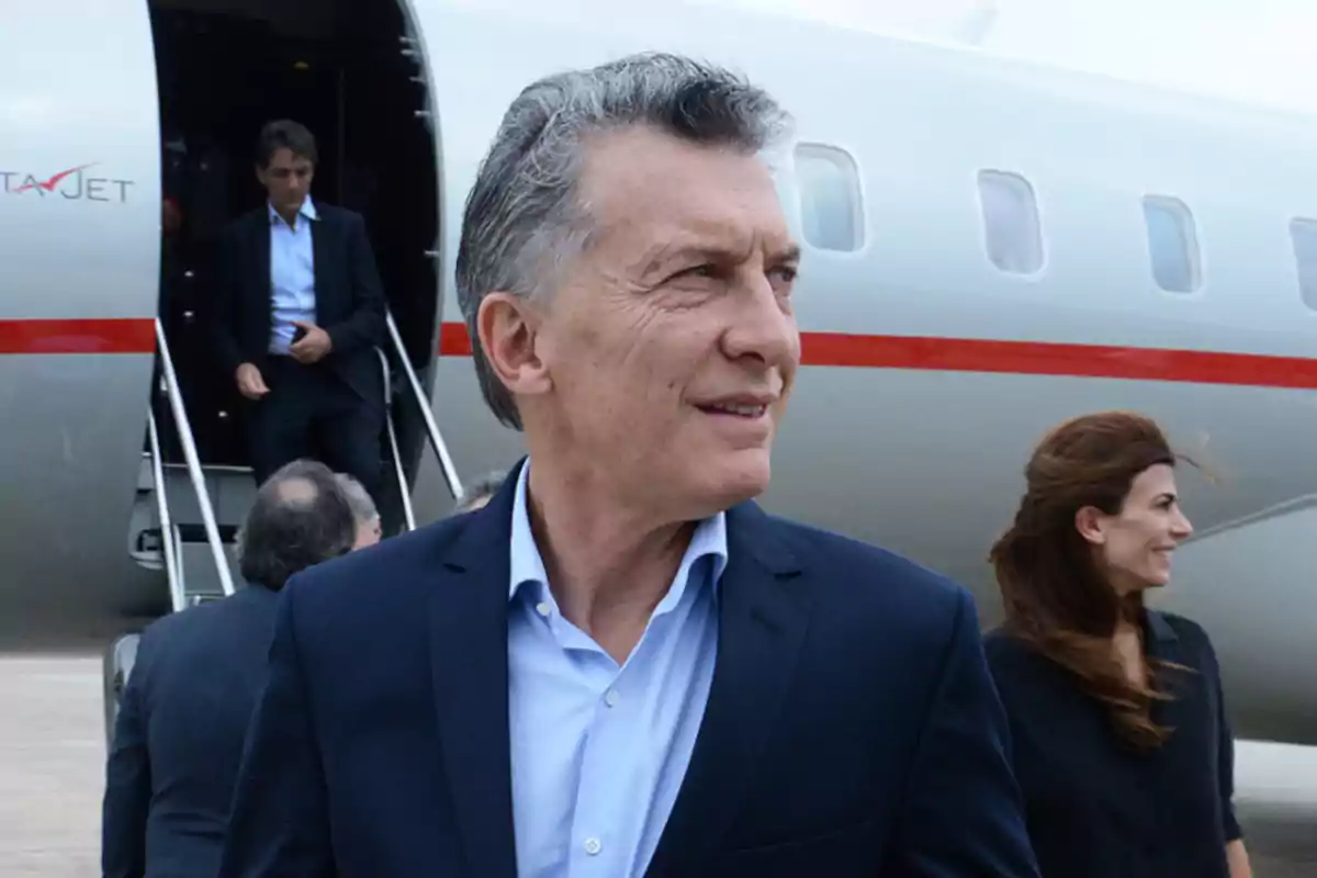 A man in a dark suit and light jersey is standing in front of a private jet, with other people descending the stairs behind him.