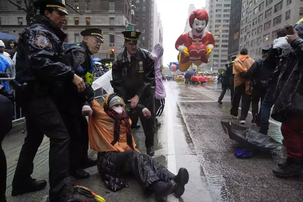 Un grupo de policías detiene a una persona en una calle mojada durante un desfile con un globo gigante de un personaje en el fondo.