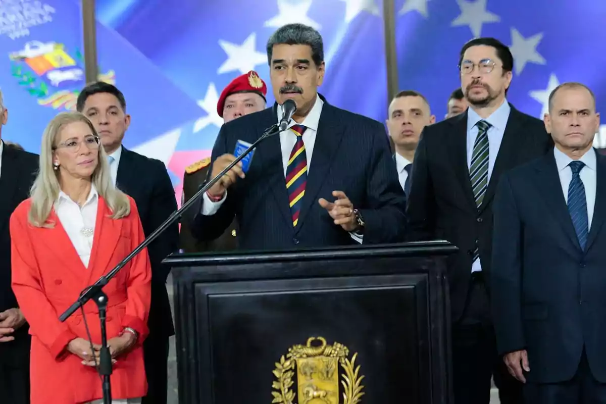 Un grupo de personas, algunas de ellas con trajes formales, se encuentran de pie detrás de un podio con un escudo, mientras una persona en el centro habla por un micrófono.