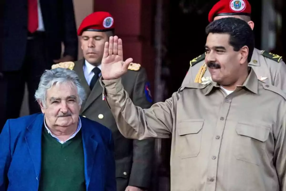 Two men in the foreground, one of them waving with his hand raised while the other stands beside him; in the background, there are two people wearing red berets and military uniforms. Two men in the foreground, one of them waving with his hand raised while the other stands beside him; in the background, there are two people wearing red berets and military uniforms.
