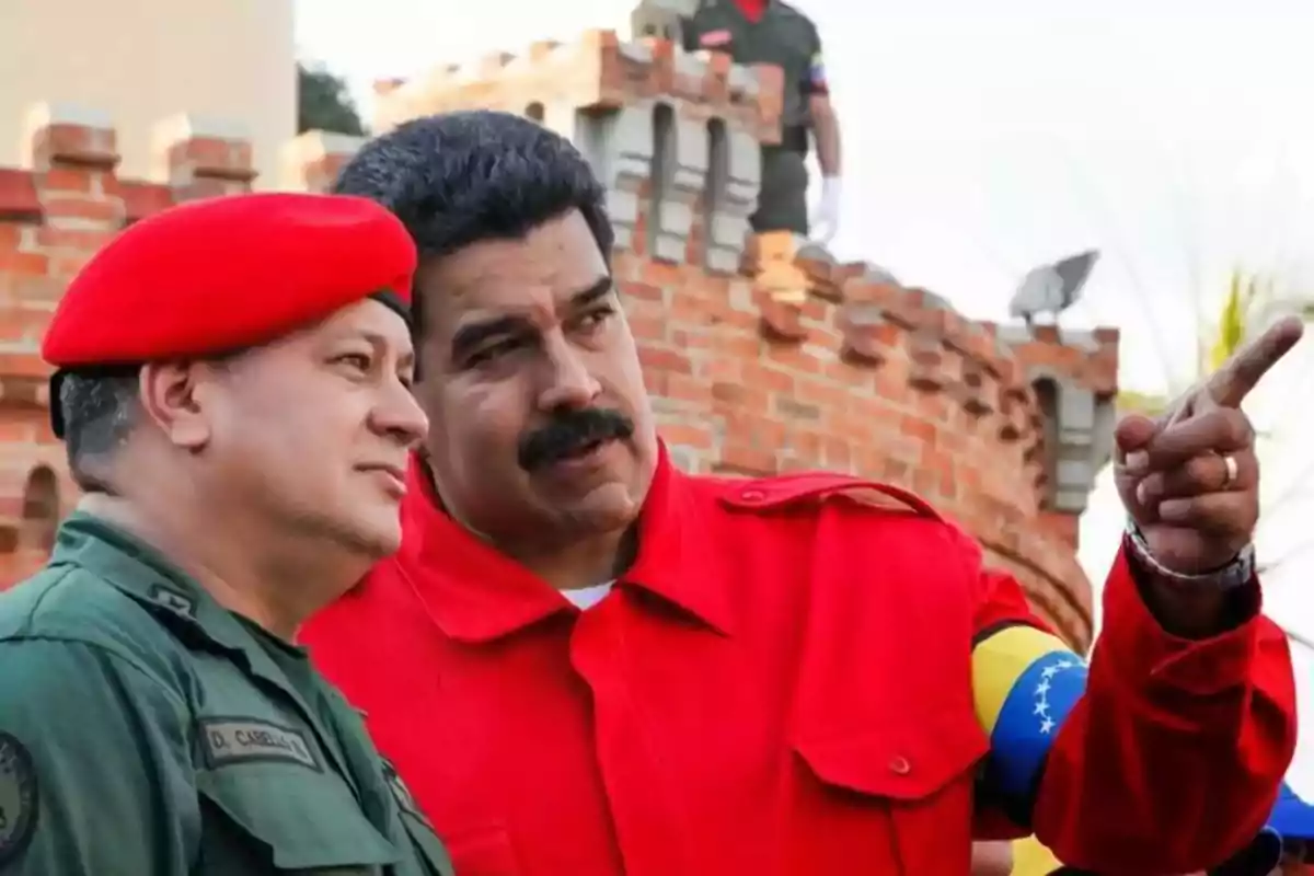 Dos hombres con uniforme militar y chaqueta roja conversan frente a una estructura de ladrillo.