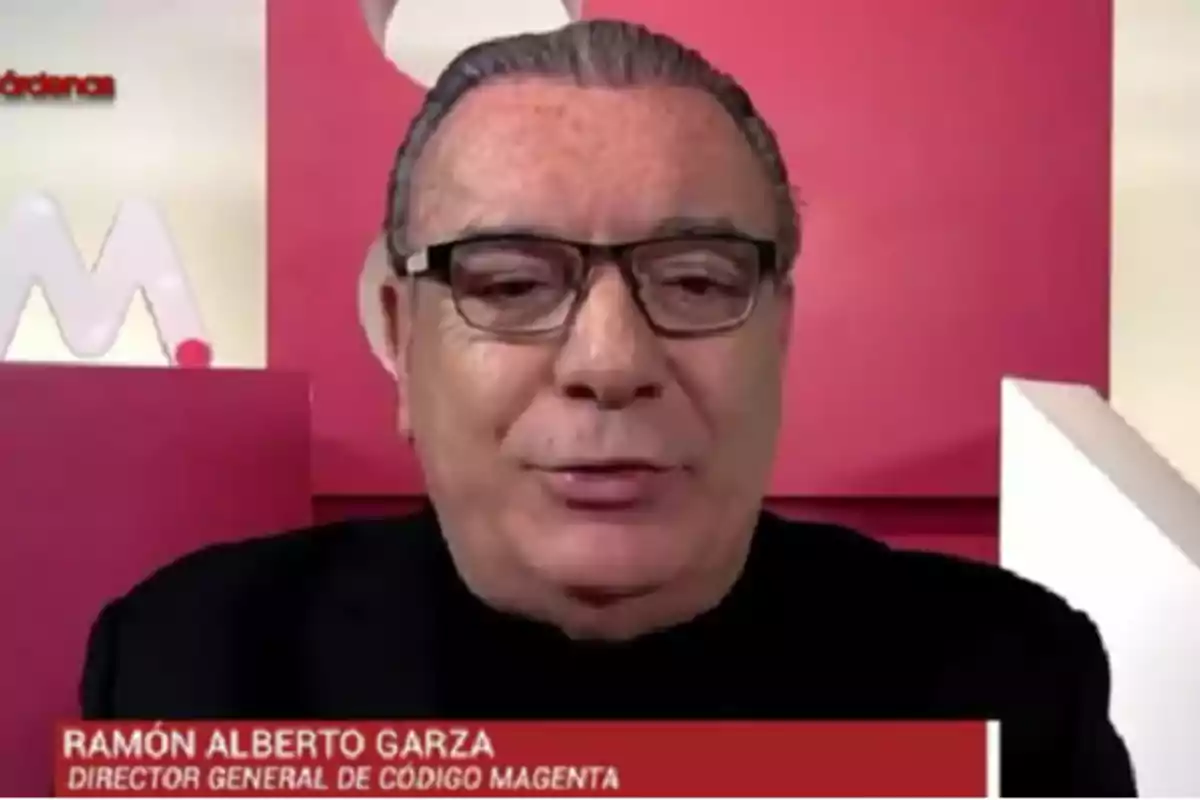 A man with glasses and slicked-back hair appears in a broadcast with a red and white background.