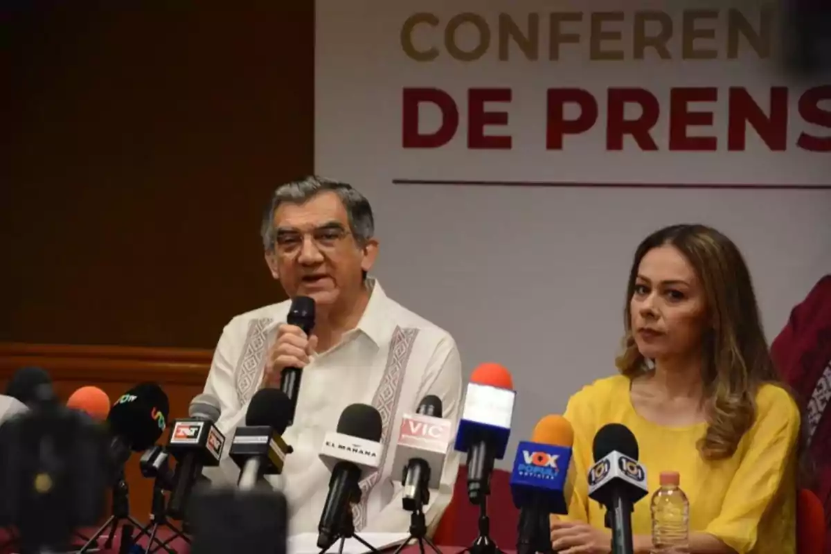 Two people at a press conference, surrounded by several microphones from different media outlets.