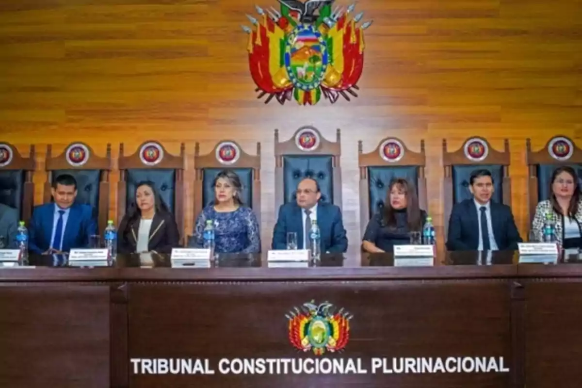Un grupo de personas sentadas en una mesa de madera con el escudo de Bolivia en la pared detrás de ellos. Un grupo de personas sentadas en una mesa de madera con el escudo de Bolivia en la pared detrás de ellos.