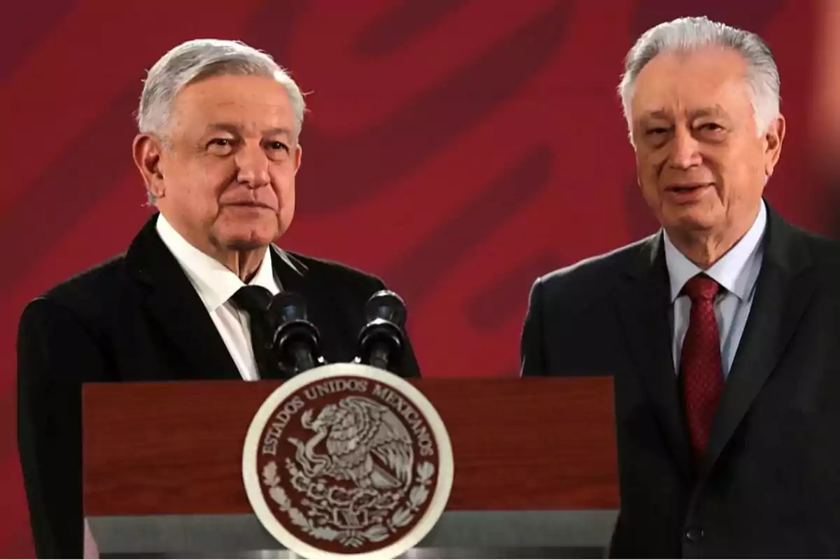 Dos hombres de pie detrás de un podio con el escudo de México, en un fondo rojo.