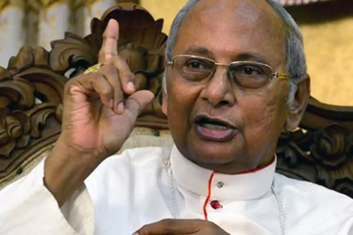 An elderly man with glasses and white religious attire is sitting on a carved chair, raising a finger as he speaks. An elderly man with glasses and white religious attire is sitting on a carved chair, raising a finger as he speaks.