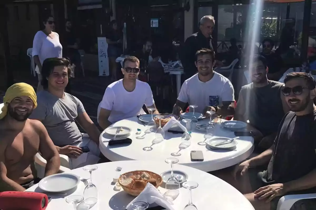 Six men smile while sitting around a round table outdoors at a restaurant, with empty plates and glasses, enjoying a sunny day. Six men smile while sitting around a round table outdoors at a restaurant, with empty plates and glasses, enjoying a sunny day.