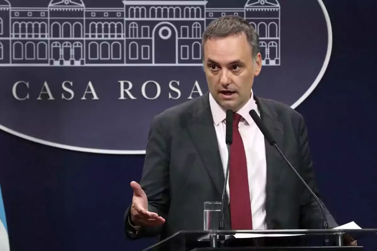 A man in a dark suit and red tie speaks at a podium in front of a background that says A man in a dark suit and red tie speaks at a podium in front of a background that says