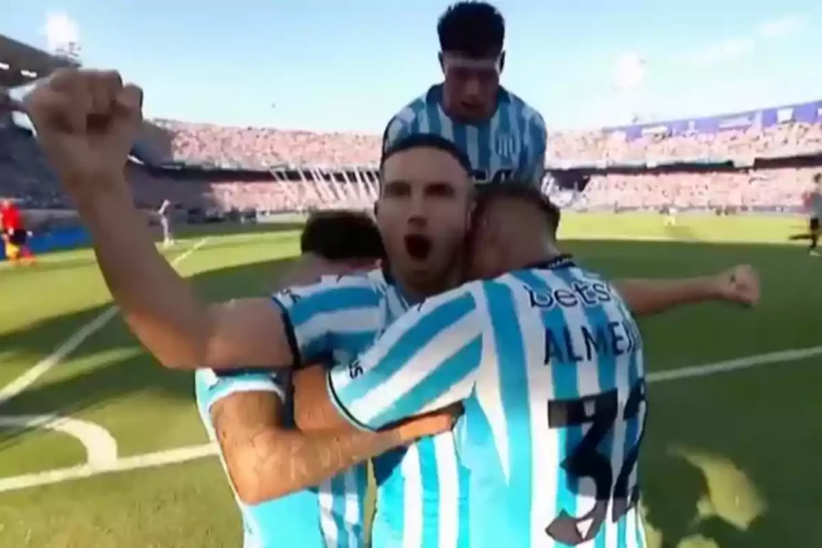 Jugadores de fútbol celebrando un gol en el campo con camisetas a rayas celestes y blancas.