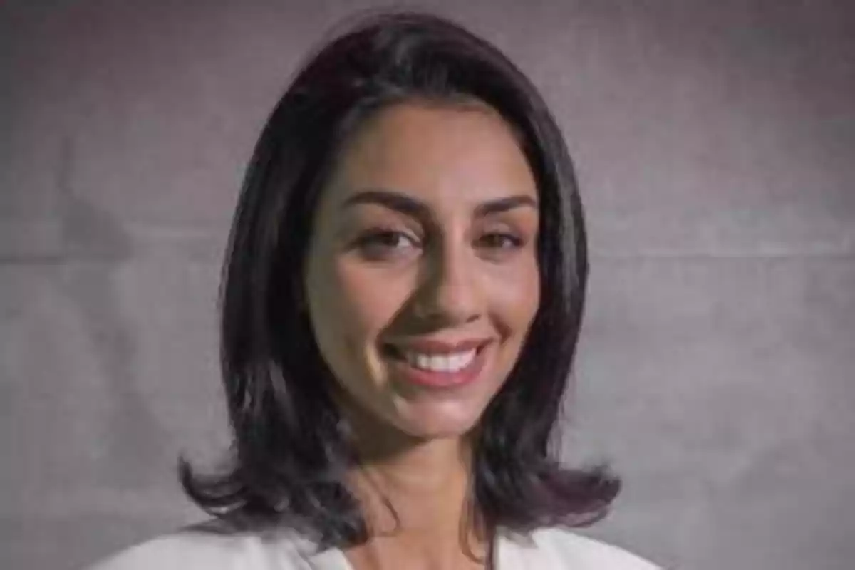 Mujer sonriendo con cabello oscuro y liso frente a un fondo gris.