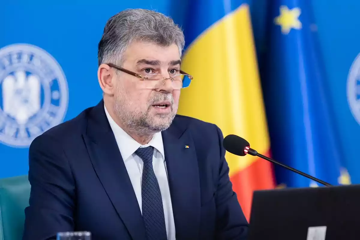 A man with a beard and glasses speaks into a microphone, wearing a dark suit and tie, with flags in the background.
