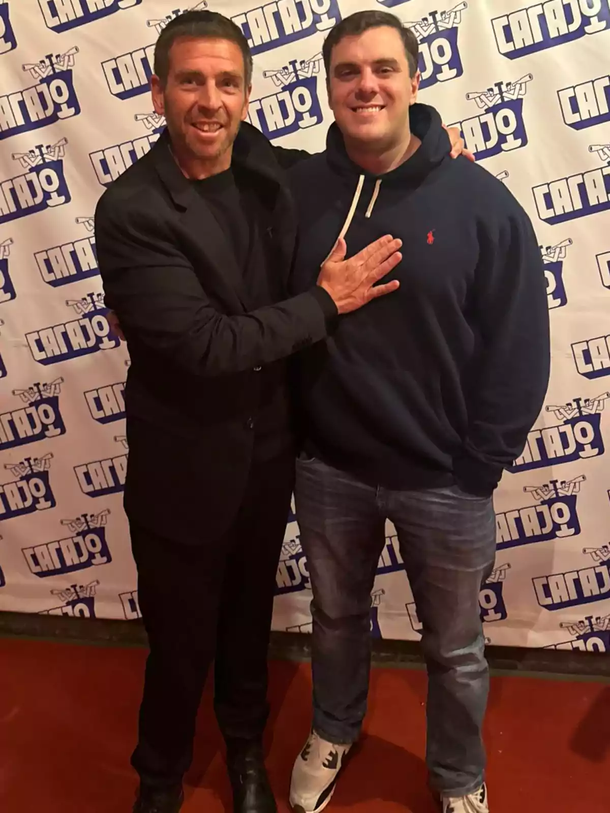 Dos hombres posan sonrientes frente a un fondo con logotipos repetidos mientras uno de ellos apoya la mano en el pecho del otro Dos hombres posan sonrientes frente a un fondo con logotipos repetidos mientras uno de ellos apoya la mano en el pecho del otro