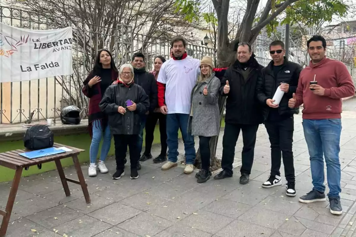 Un grupo de personas posando sonrientes en la vereda junto a una mesa de madera y una bandera de La Libertad Avanza en La Falda, algunos levantan el pulgar y otros sostienen mates y termos, con árboles y una reja de fondo.
