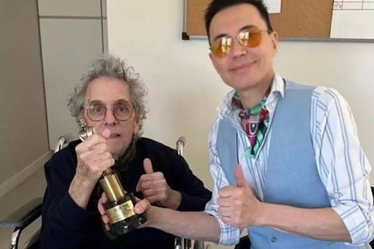 Dos personas posan juntas mientras una de ellas sostiene un trofeo y ambas muestran gestos de aprobación con el pulgar hacia arriba. Dos personas posan juntas mientras una de ellas sostiene un trofeo y ambas muestran gestos de aprobación con el pulgar hacia arriba.