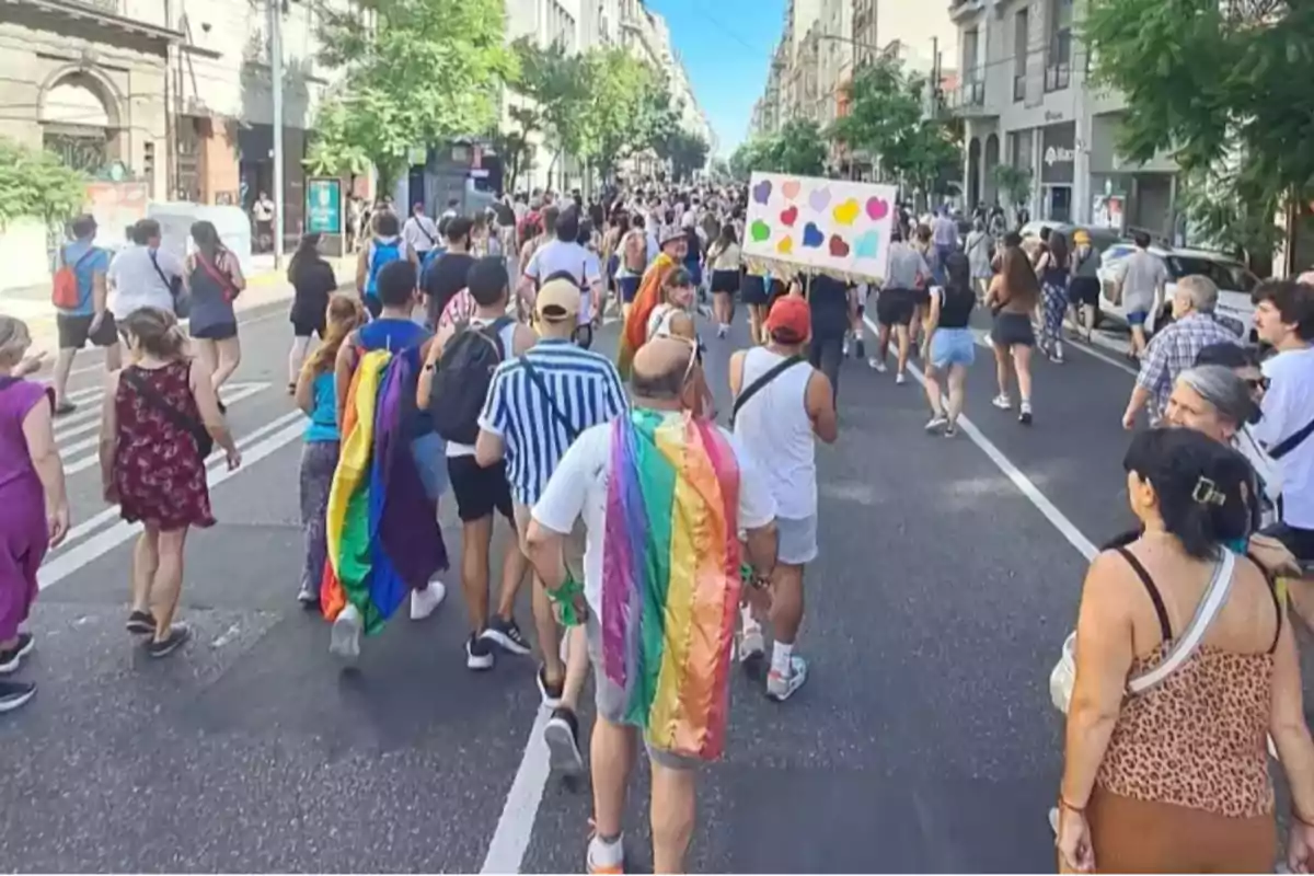 Un grupo de personas participa en una marcha en la calle, algunas llevan banderas de arcoíris y una persona sostiene un cartel con corazones de colores.