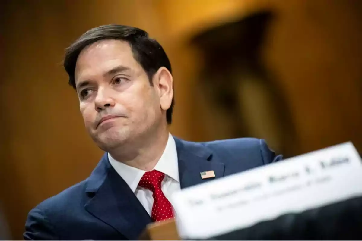 A man in a blue suit and red tie is sitting in a room, with a serious expression and a nameplate in front of him. A man in a blue suit and red tie is sitting in a room, with a serious expression and a nameplate in front of him.