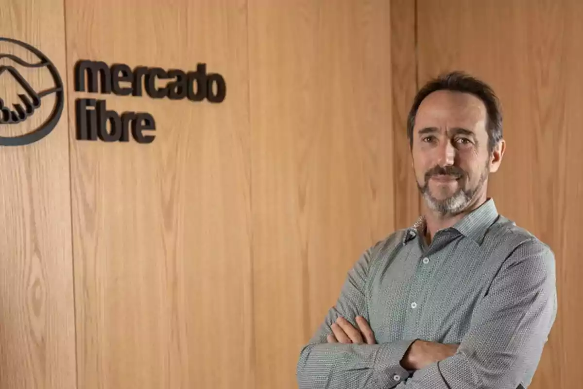 Hombre de cabello corto y barba con camisa gris de manga larga posando con los brazos cruzados frente a una pared de madera con el logo de Mercado Libre