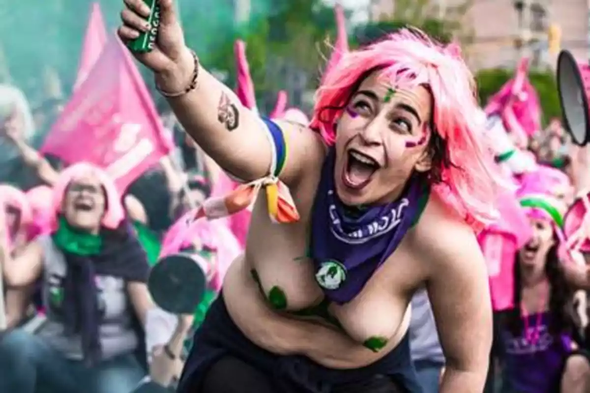 Una persona con peluca rosa y pañuelo morado participa con entusiasmo en una manifestación rodeada de otras personas con banderas y pelucas de colores.