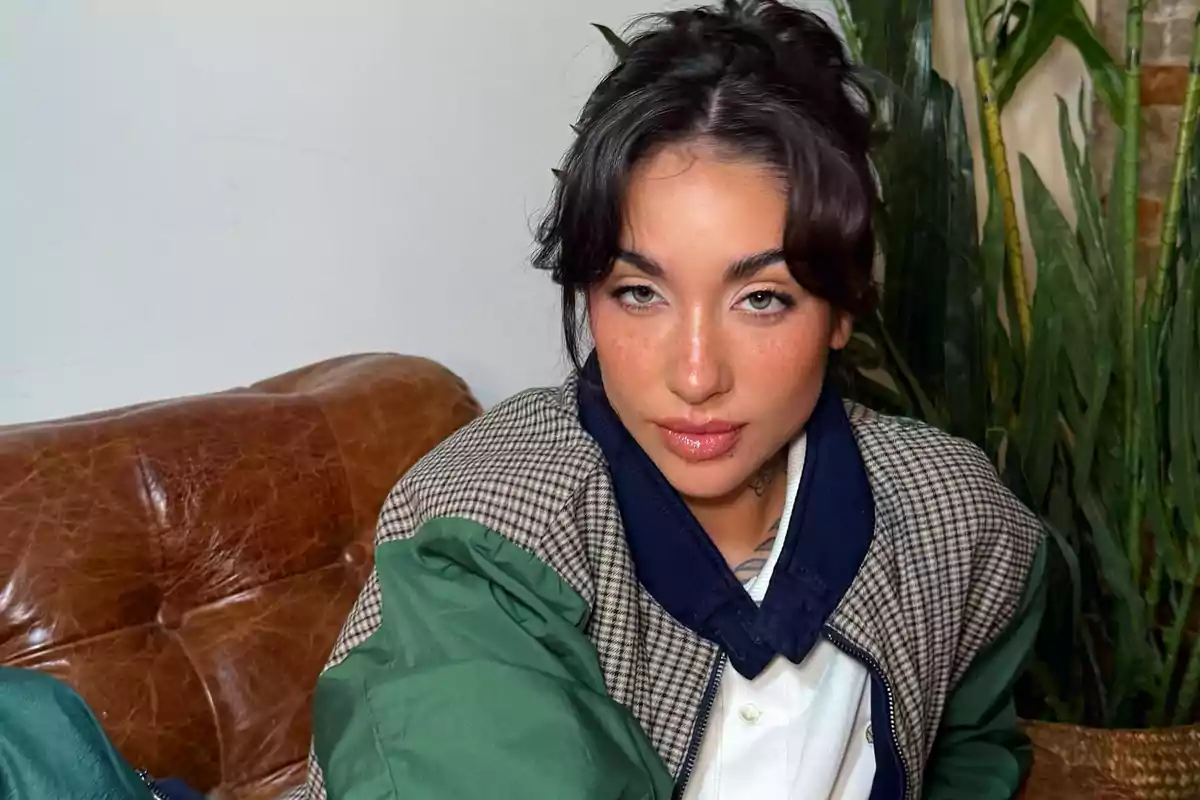 Una persona con chaqueta verde y camisa blanca está sentada en un sofá de cuero marrón con plantas al fondo.