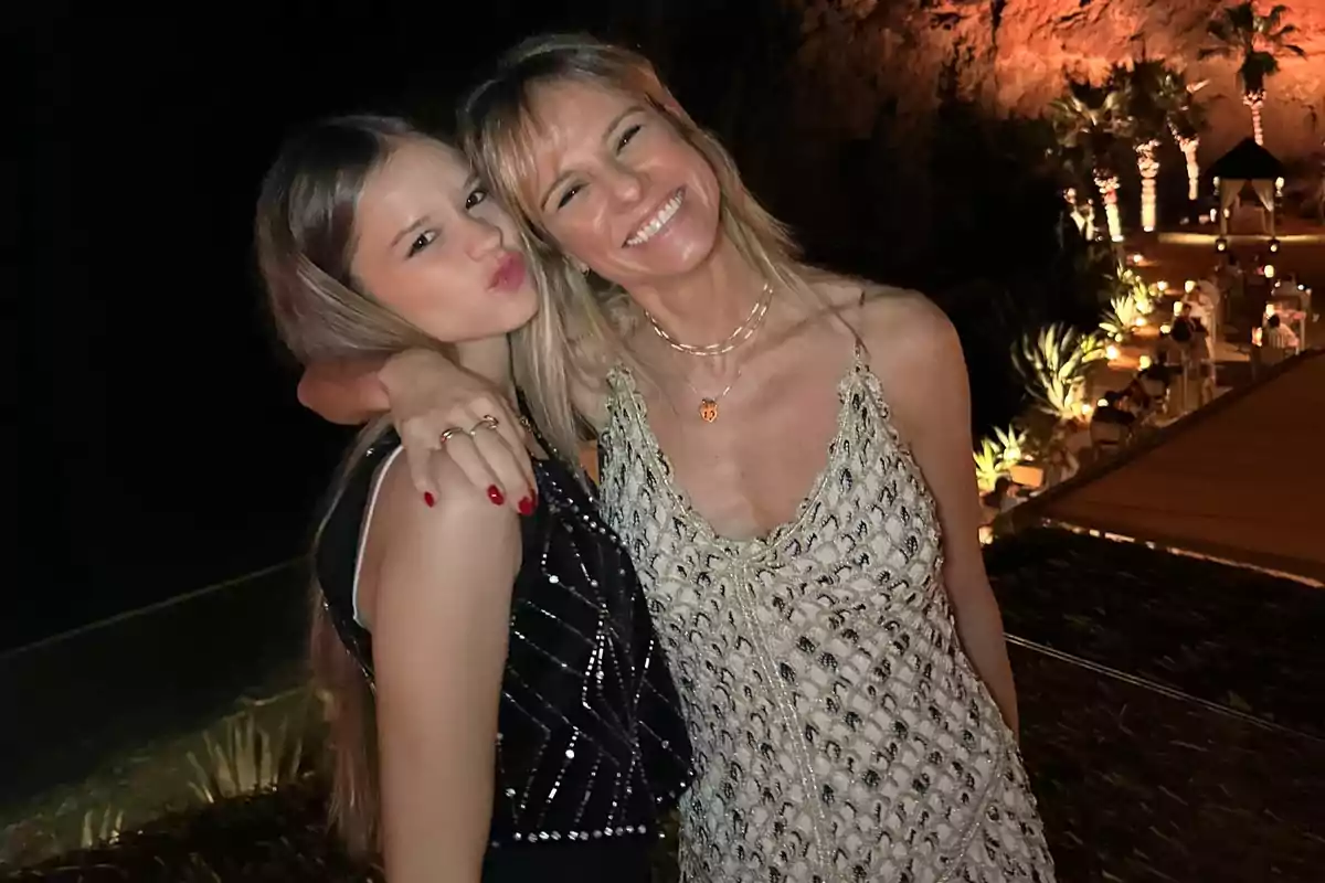 Dos personas posan juntas sonriendo en un ambiente nocturno con luces y vegetación de fondo.