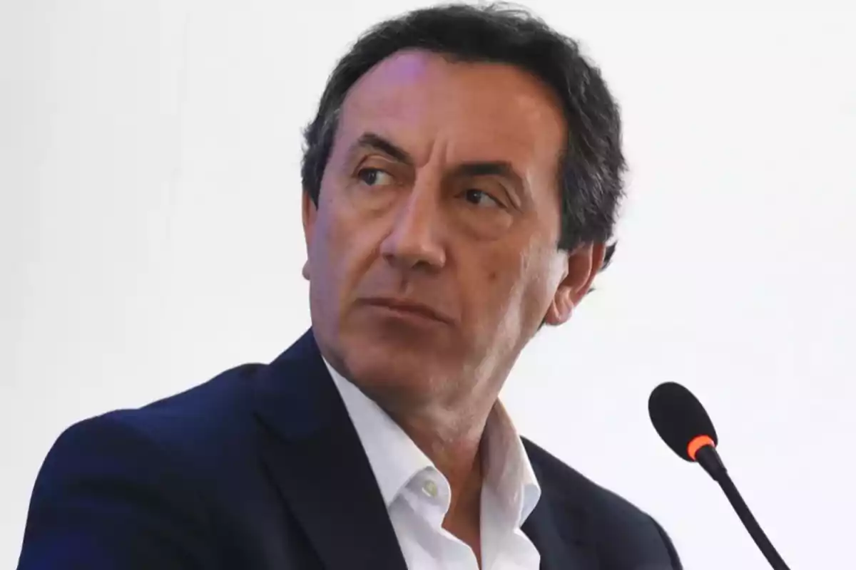 Un hombre con traje oscuro y camisa blanca está sentado frente a un micrófono mirando hacia un lado.