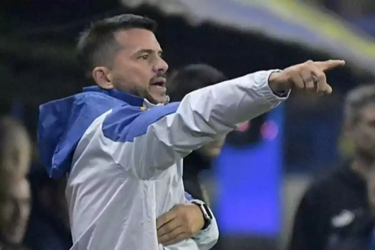 Un hombre con chaqueta deportiva blanca y azul señala con el dedo en un entorno deportivo. Un hombre con chaqueta deportiva blanca y azul señala con el dedo en un entorno deportivo.