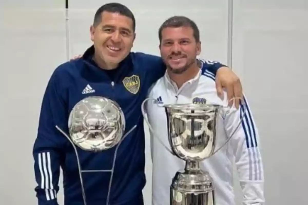 Two men smile and pose together wearing Boca Juniors sportswear while holding two silver trophies. Two men smile and pose together wearing Boca Juniors sportswear while holding two silver trophies.