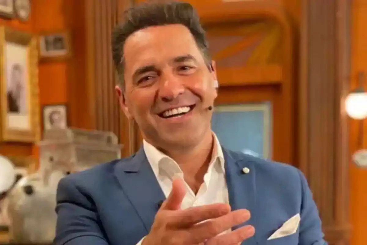 Un hombre sonriente con un micrófono en la oreja, vistiendo un traje azul, en un ambiente interior con decoración de madera y cuadros en la pared. Un hombre sonriente con un micrófono en la oreja, vistiendo un traje azul, en un ambiente interior con decoración de madera y cuadros en la pared.