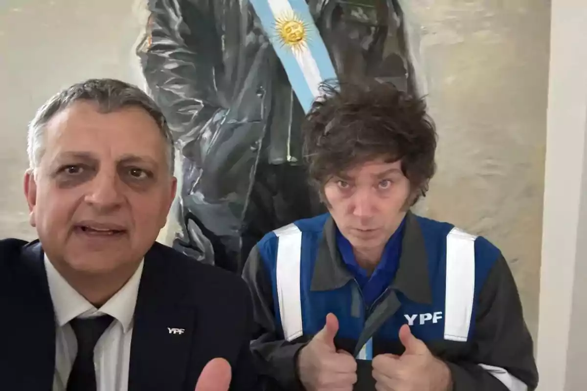 Two people pose for a photo, one of them wearing a formal suit and the other dressed in a YPF uniform; both are standing in front of a painting with a light blue and white stripe.