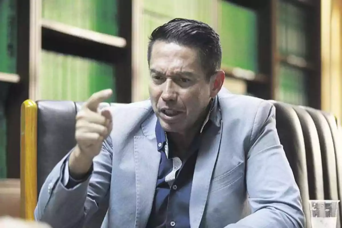 Hombre con expresión seria y gesto enfático sentado en una silla con estanterías de libros al fondo.