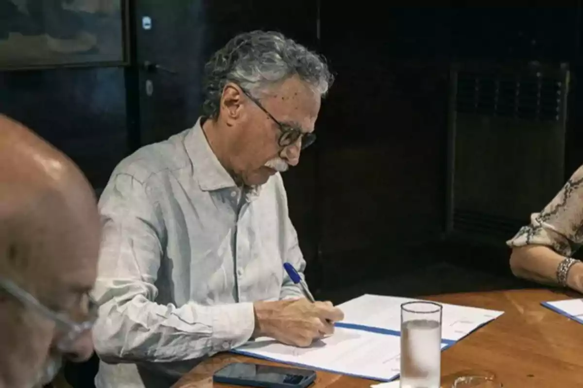 Un hombre de cabello canoso y gafas está sentado en una mesa firmando documentos, con un vaso de agua y un teléfono móvil cerca.