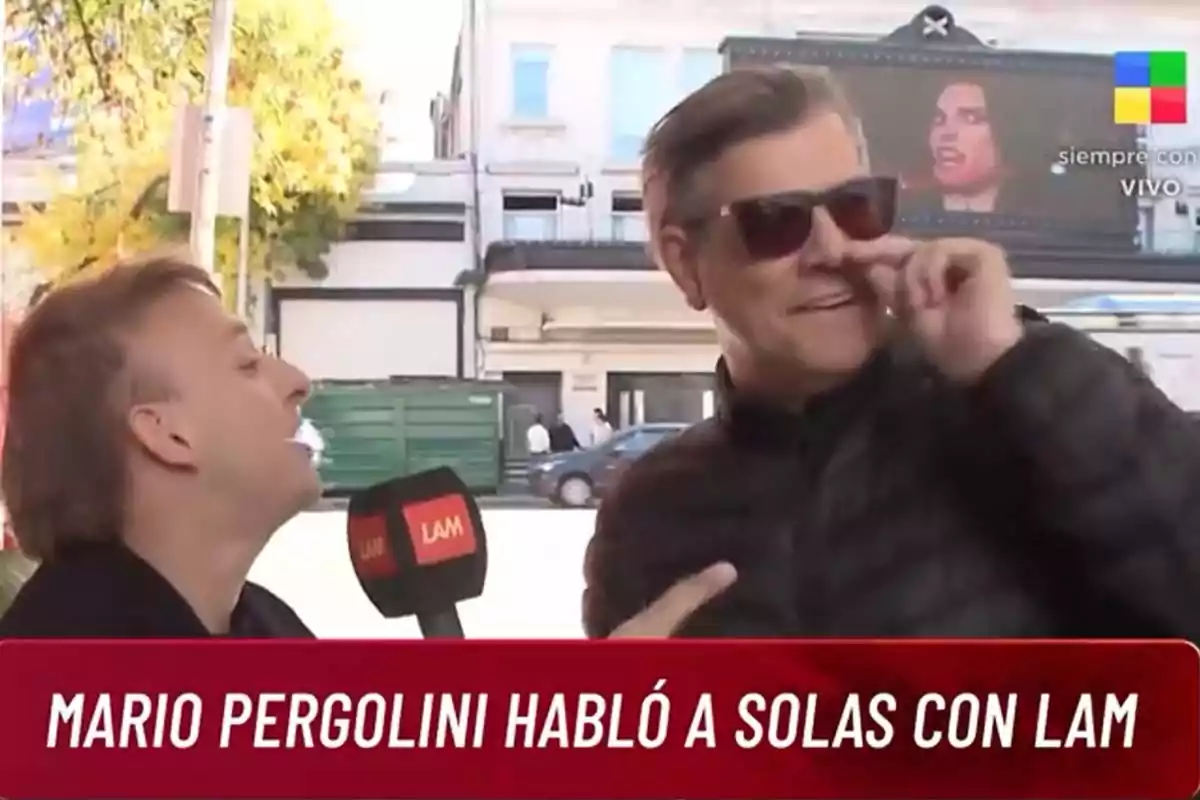 Dos personas están en una entrevista al aire libre, una de ellas sostiene un micrófono con el logo de LAM, mientras la otra persona se toca la nariz y lleva gafas de sol. Dos personas están en una entrevista al aire libre, una de ellas sostiene un micrófono con el logo de LAM, mientras la otra persona se toca la nariz y lleva gafas de sol.