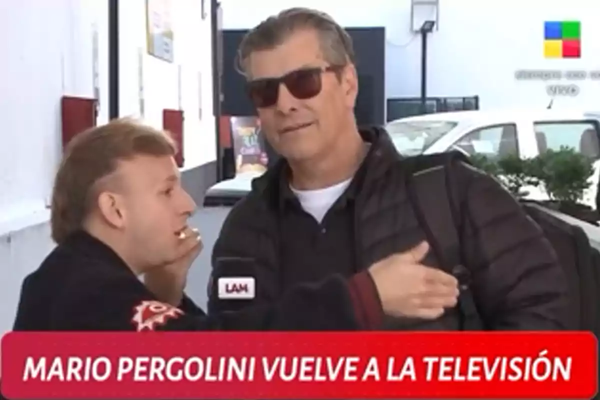 Dos personas conversando en la calle, una de ellas sostiene un micrófono con el logo Dos personas conversando en la calle, una de ellas sostiene un micrófono con el logo