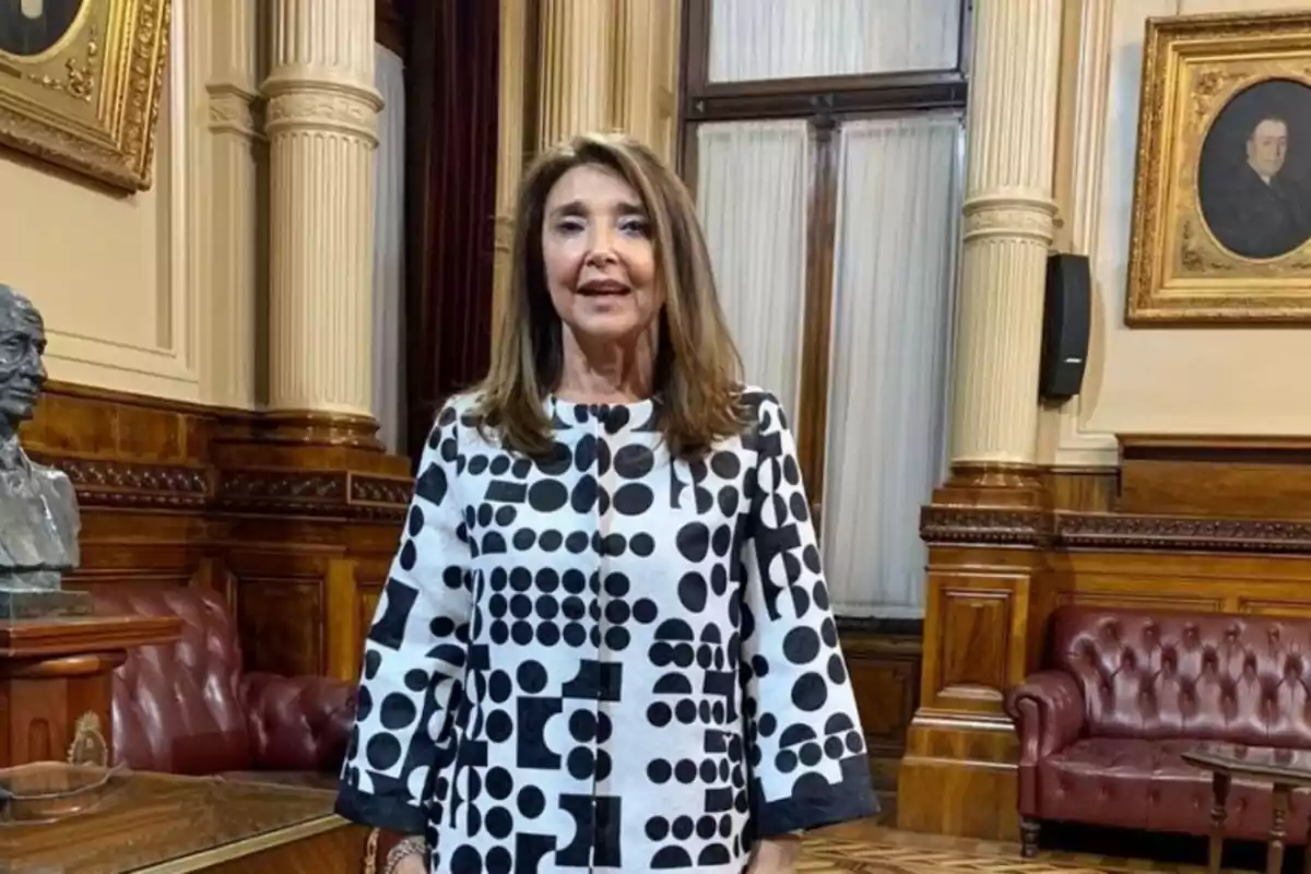 Mujer de cabello castaño y liso con saco blanco de estampado negro de círculos y cuadrados, posando en una sala elegante con sillones de cuero, columnas y retratos enmarcados en la pared. Mujer de cabello castaño y liso con saco blanco de estampado negro de círculos y cuadrados, posando en una sala elegante con sillones de cuero, columnas y retratos enmarcados en la pared.