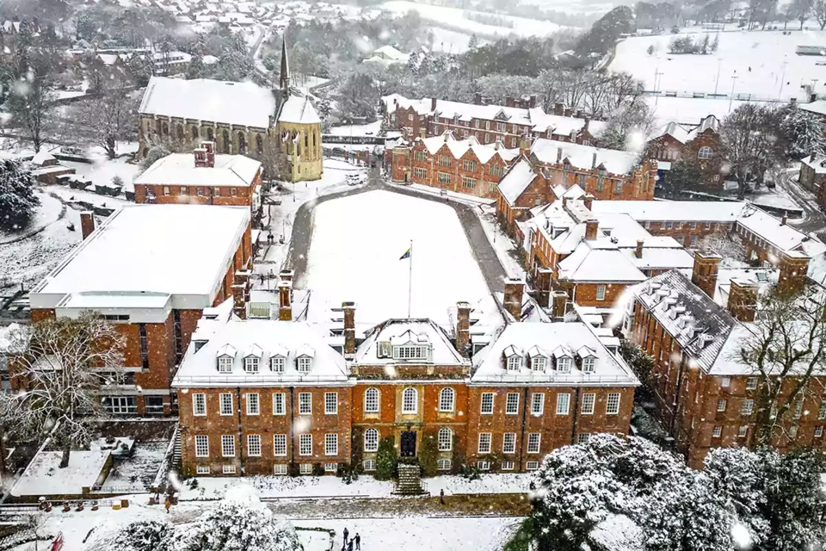 Vista aérea de un campus universitario cubierto de nieve, con edificios de ladrillo rojo y una iglesia con torre en el centro. Vista aérea de un campus universitario cubierto de nieve, con edificios de ladrillo rojo y una iglesia con torre en el centro.