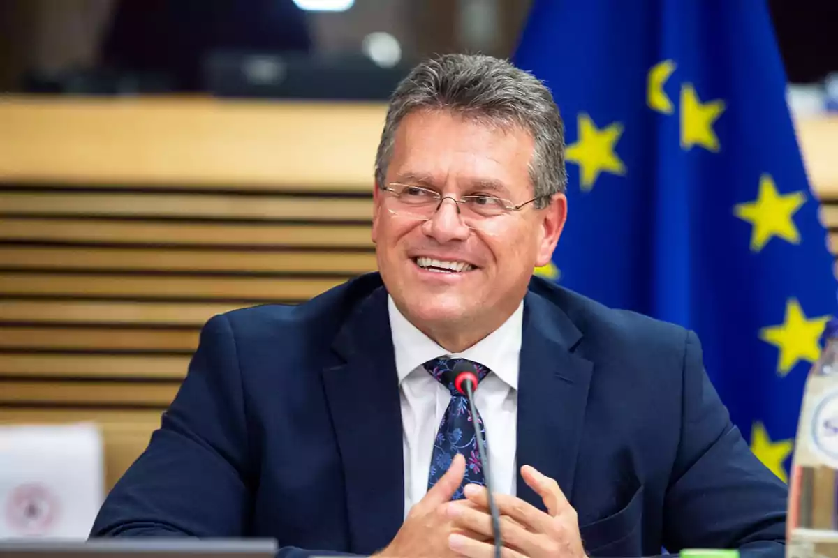 Un hombre con gafas y traje oscuro sonríe mientras está sentado frente a un micrófono con una bandera de la Unión Europea de fondo. Un hombre con gafas y traje oscuro sonríe mientras está sentado frente a un micrófono con una bandera de la Unión Europea de fondo.