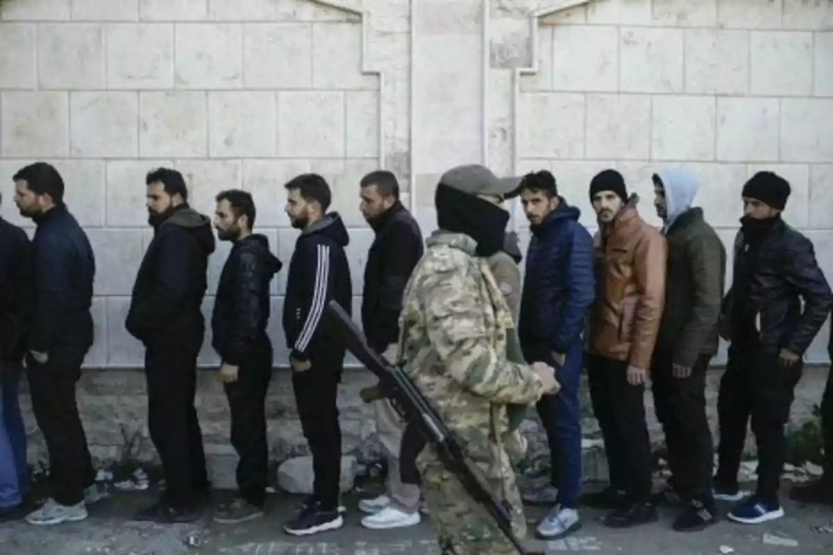 A group of people stands in line next to a wall while an armed individual with a covered face walks in front of them.