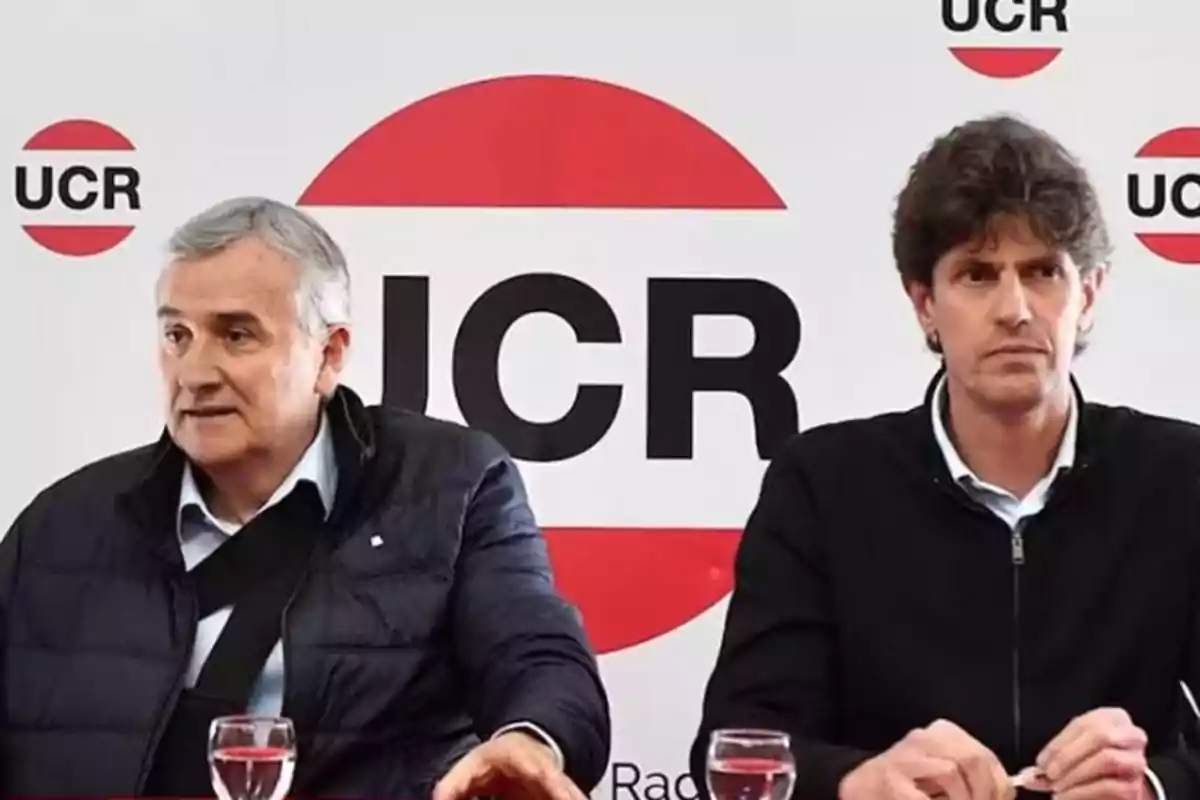 Dos personas sentadas frente a un fondo con el logotipo de la UCR.