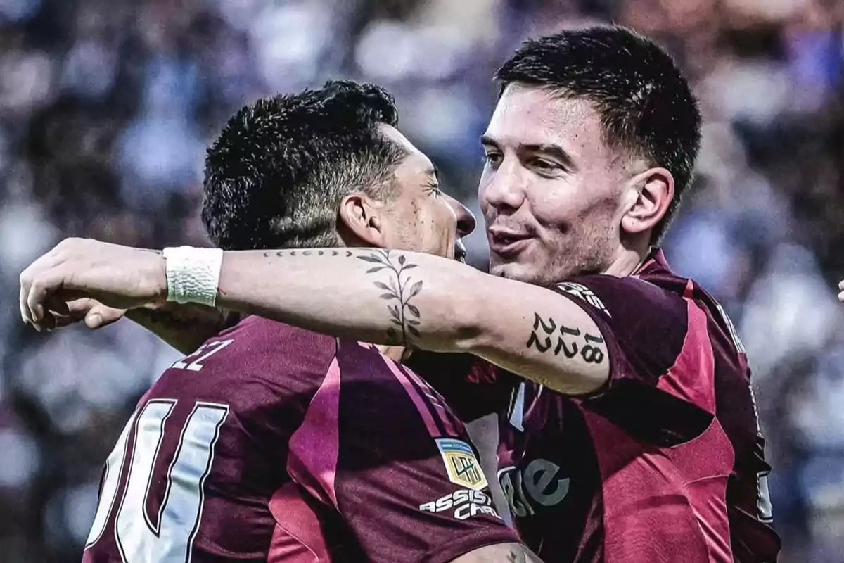 Two players in maroon uniforms hug each other celebrating a goal in a packed stadium. Two players in maroon uniforms hug each other celebrating a goal in a packed stadium.