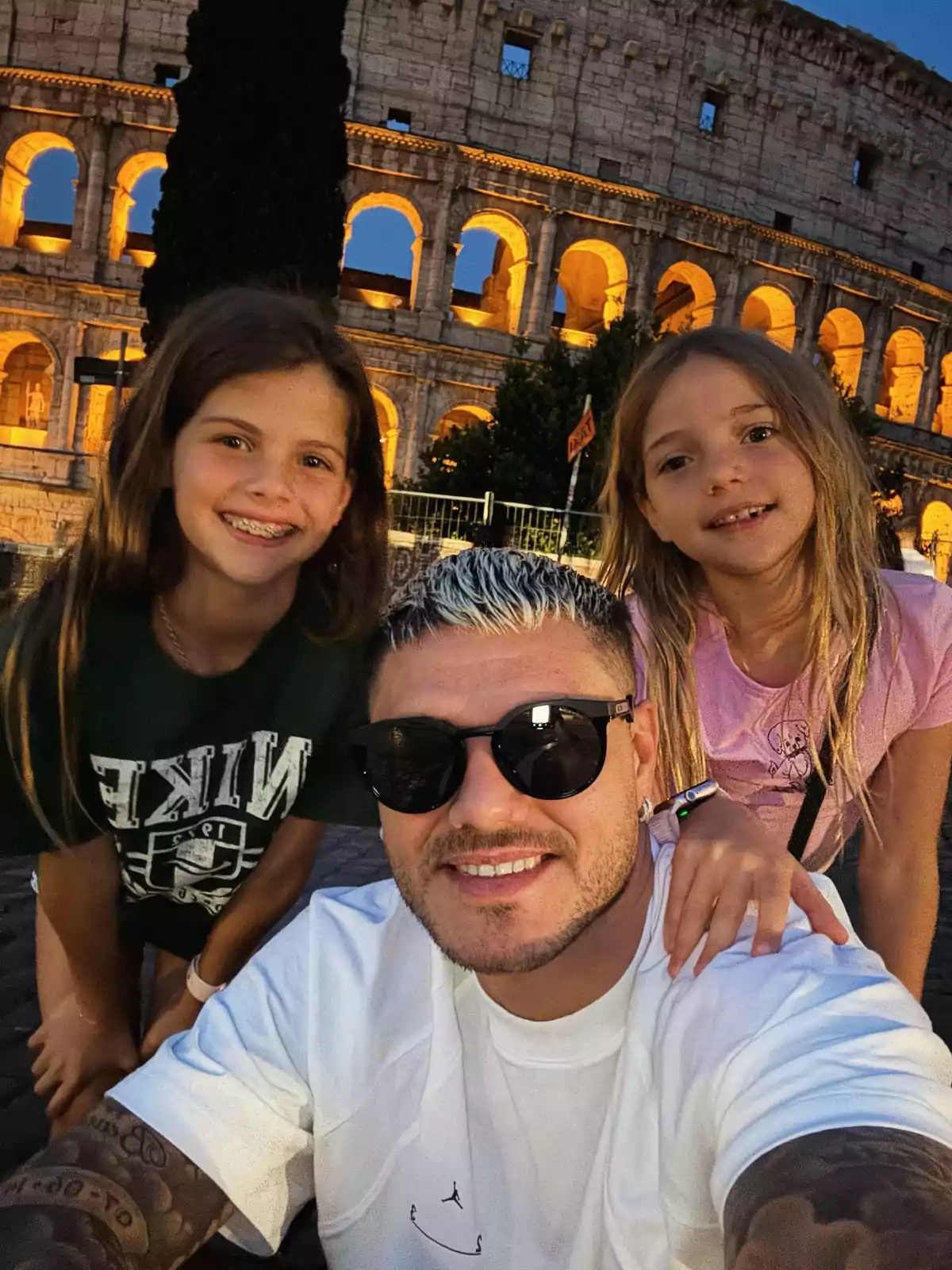 A man with sunglasses and two girls smile in front of the Coliseum lit up at sunset. A man with sunglasses and two girls smile in front of the Coliseum lit up at sunset.