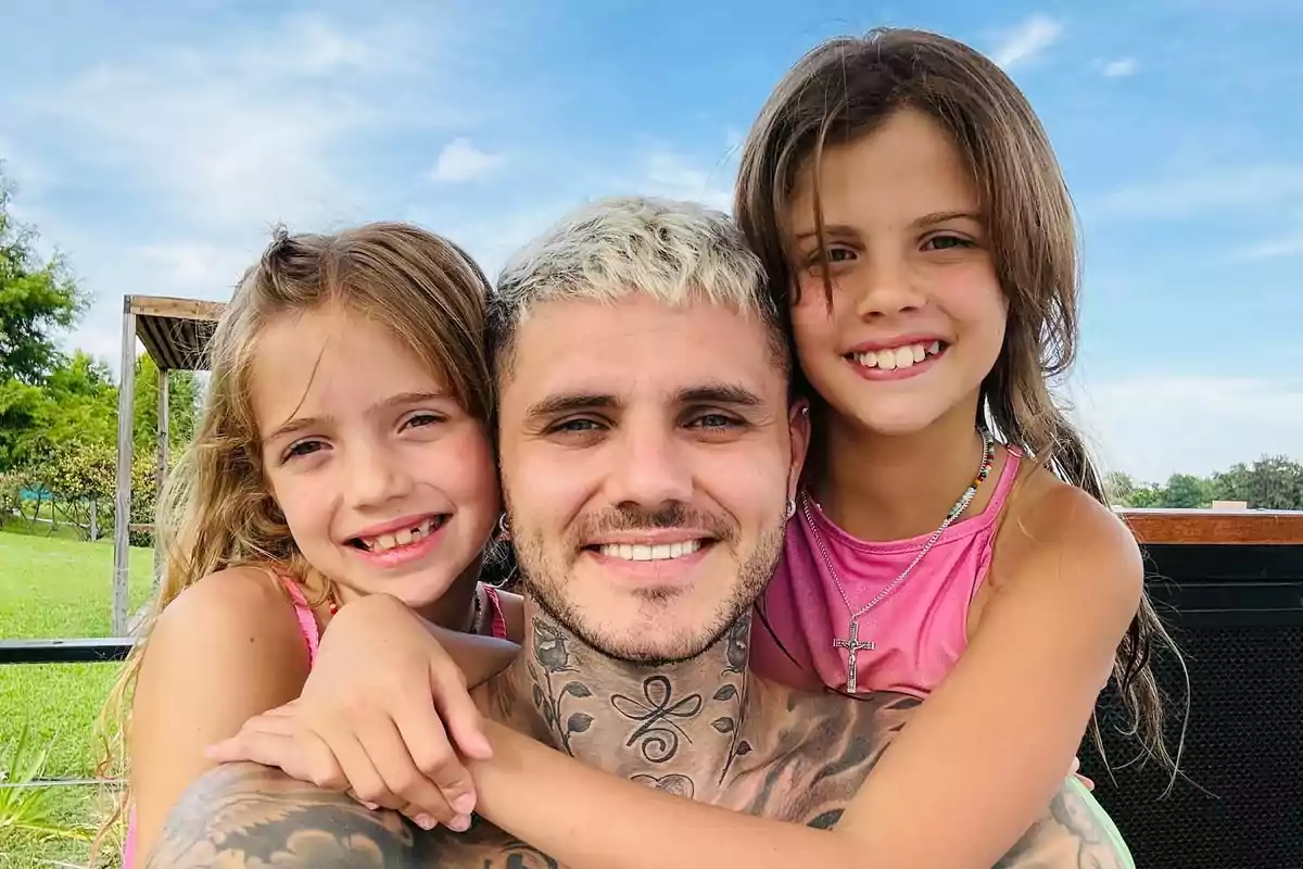 A smiling man with short blond hair poses with his arms around two little girls who are also smiling, all of them outdoors on a sunny day. A smiling man with short blond hair poses with his arms around two little girls who are also smiling, all of them outdoors on a sunny day.