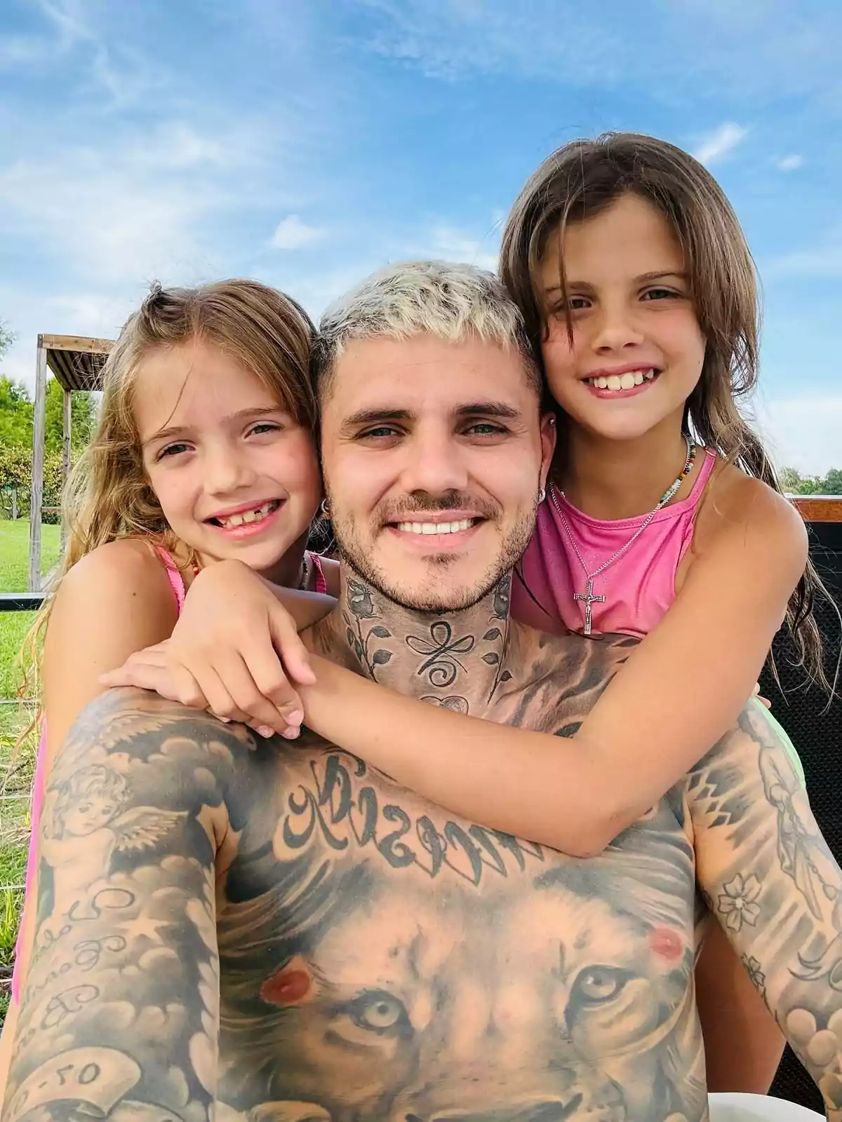 Un hombre con tatuajes en el pecho y el cuello sonríe mientras dos niñas lo abrazan por los hombros, todos están al aire libre bajo un cielo azul. Un hombre con tatuajes en el pecho y el cuello sonríe mientras dos niñas lo abrazan por los hombros, todos están al aire libre bajo un cielo azul.