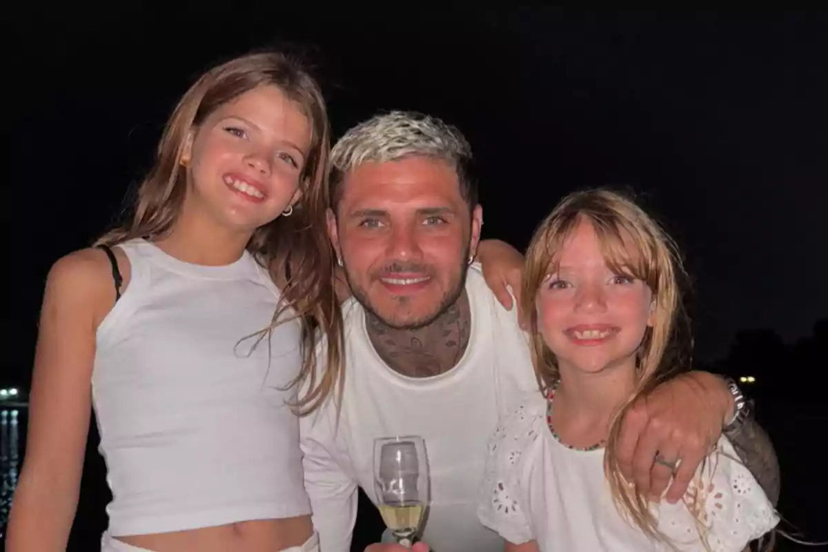 Three people smile, hugging each other in front of a dark background; one of them is holding a glass.
