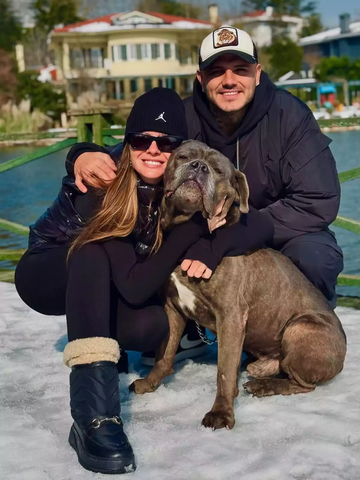 Una pareja sonriente posa junto a su perro en un entorno nevado con una casa y un lago de fondo. Una pareja sonriente posa junto a su perro en un entorno nevado con una casa y un lago de fondo.