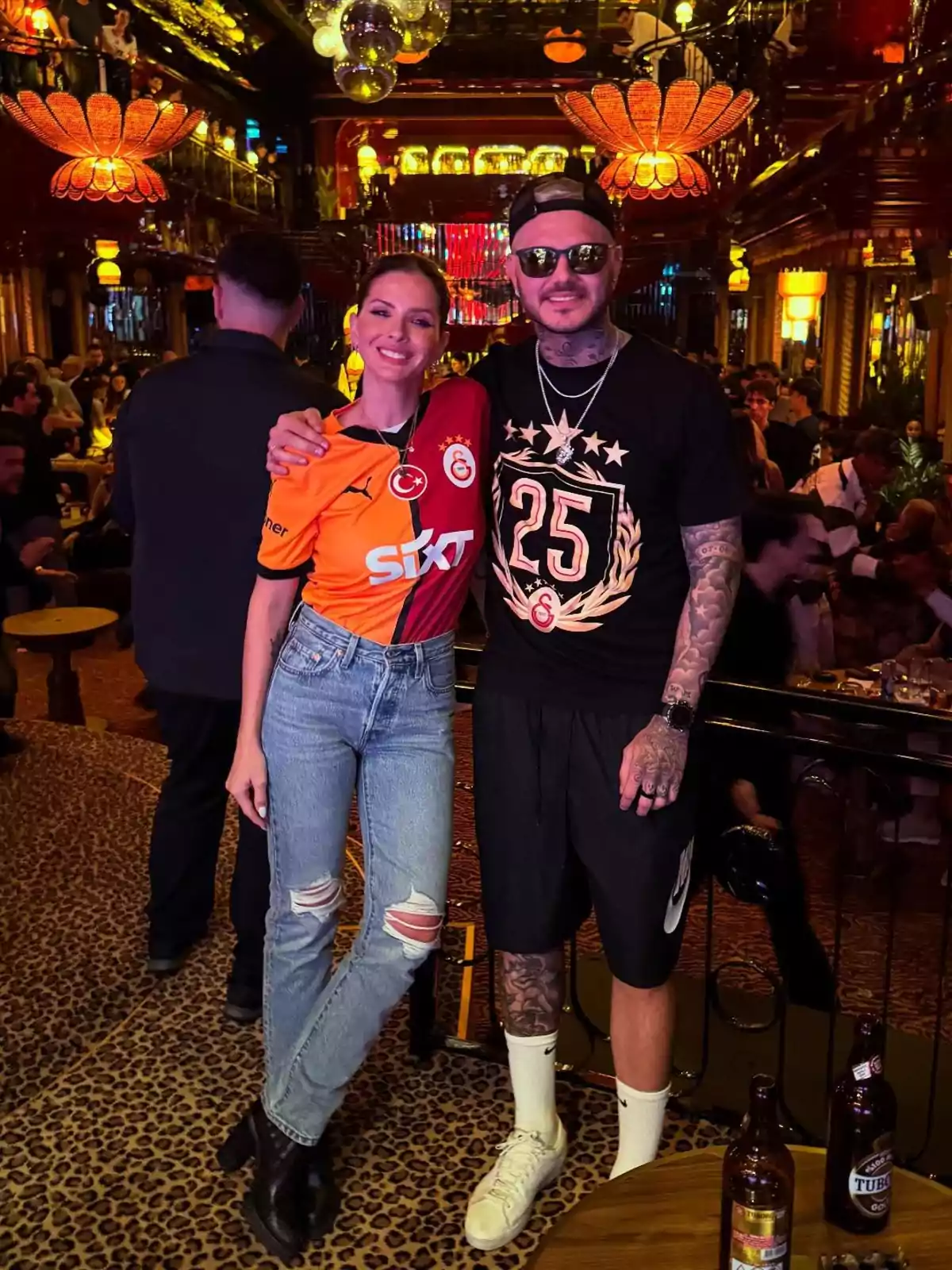 A woman and a man pose together in a bar with striking decor and warm lighting, she is wearing an orange sports jersey and he is dressed in casual clothes with visible tattoos. A woman and a man pose together in a bar with striking decor and warm lighting, she is wearing an orange sports jersey and he is dressed in casual clothes with visible tattoos.