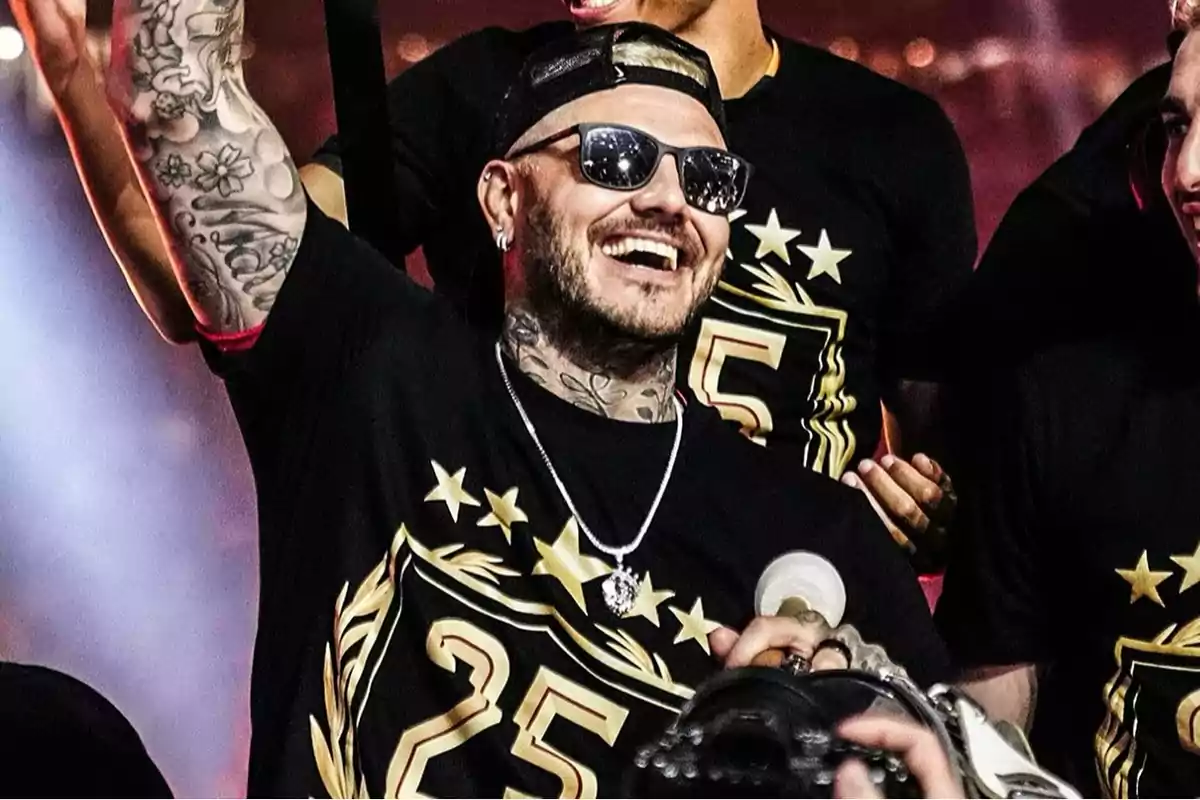 A man with sunglasses and a cap smiles while raising his arm, surrounded by people wearing black jerseys decorated with stars and the number 25.