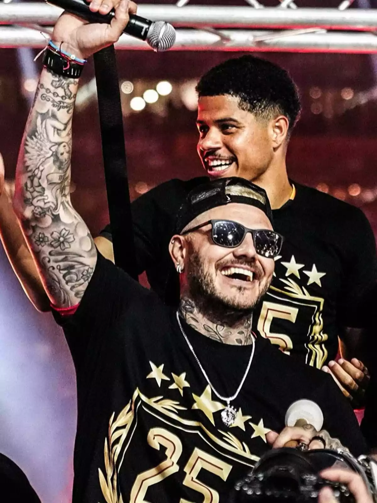 Two smiling people at an event, one of them with tattoos and sunglasses, holding up a microphone. Two smiling people at an event, one of them with tattoos and sunglasses, holding up a microphone.
