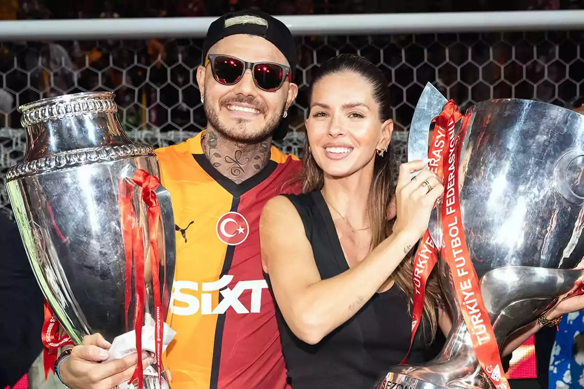 Dos personas sonríen mientras sostienen grandes trofeos plateados en un estadio de fútbol, una de ellas lleva una camiseta del Galatasaray y gafas de sol