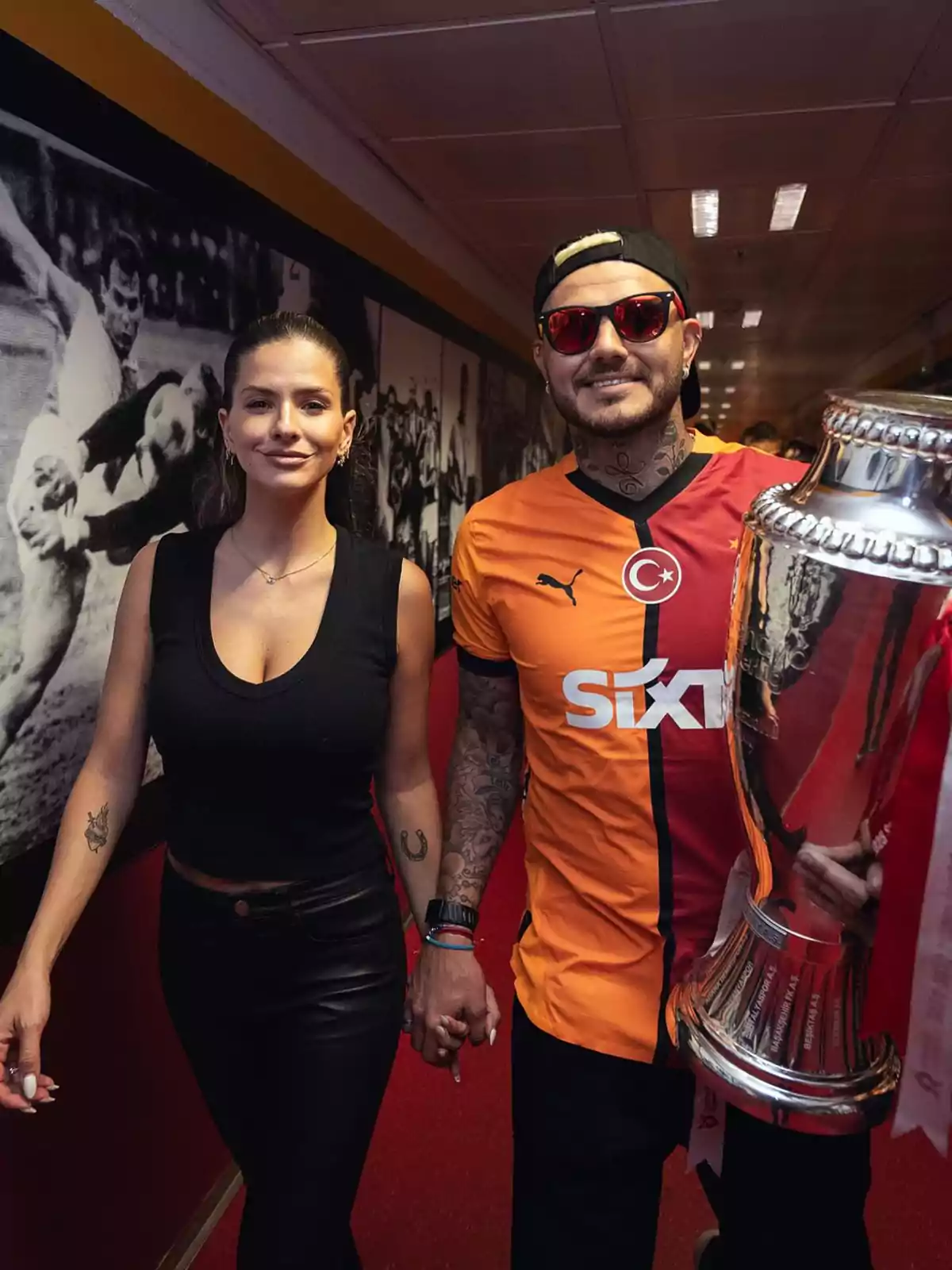 Una pareja sonriente camina tomada de la mano en un pasillo, el hombre lleva una camiseta de fútbol naranja y roja y sostiene un trofeo plateado grande, mientras la mujer viste de negro y ambos lucen tatuajes en los brazos. Una pareja sonriente camina tomada de la mano en un pasillo, el hombre lleva una camiseta de fútbol naranja y roja y sostiene un trofeo plateado grande, mientras la mujer viste de negro y ambos lucen tatuajes en los brazos.