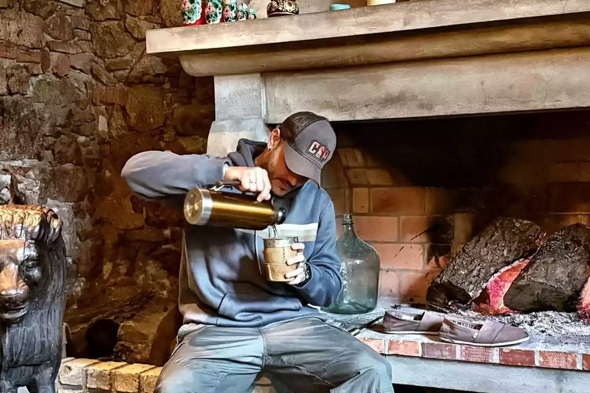 Hombre sentado frente a una chimenea encendida sirviendo una bebida caliente en un vaso con un termo mientras usa gorra y sudadera Hombre sentado frente a una chimenea encendida sirviendo una bebida caliente en un vaso con un termo mientras usa gorra y sudadera